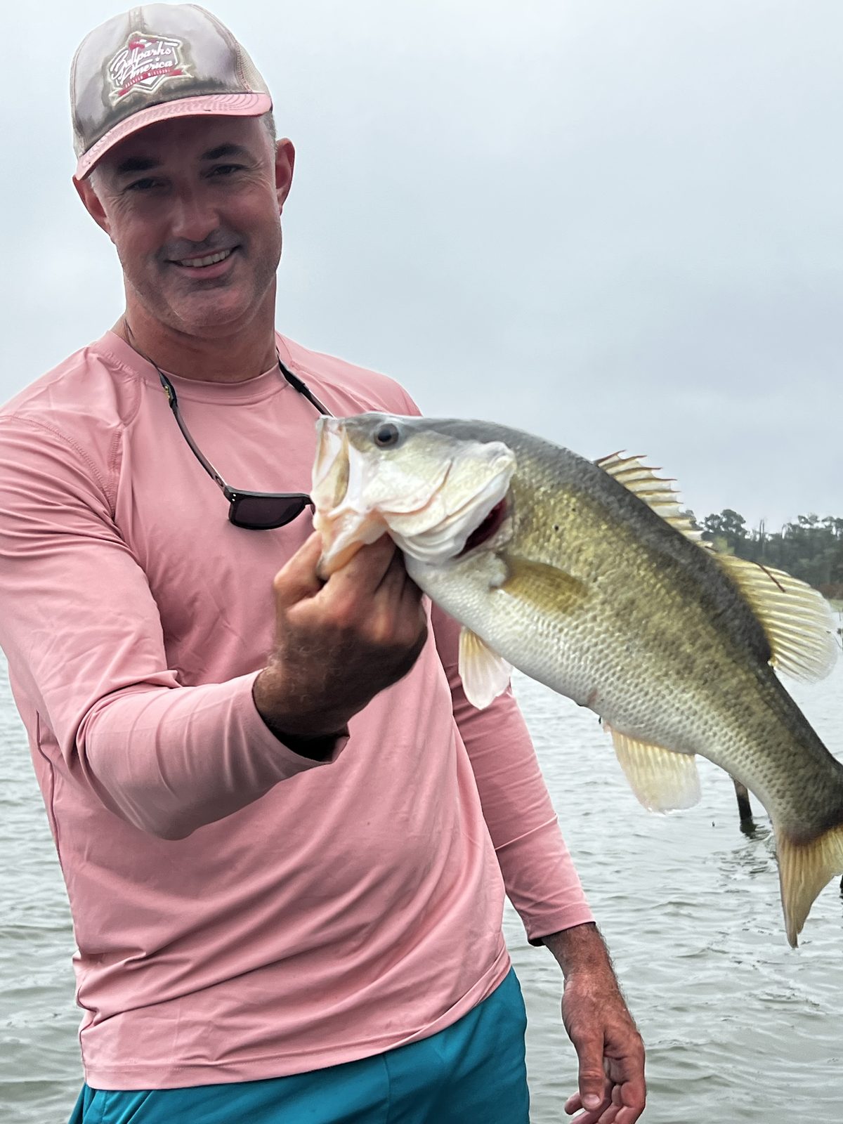 Close-up of Jeremiah King in a pink sun shirt and cap holding a big largemouth bass on an overcast lake