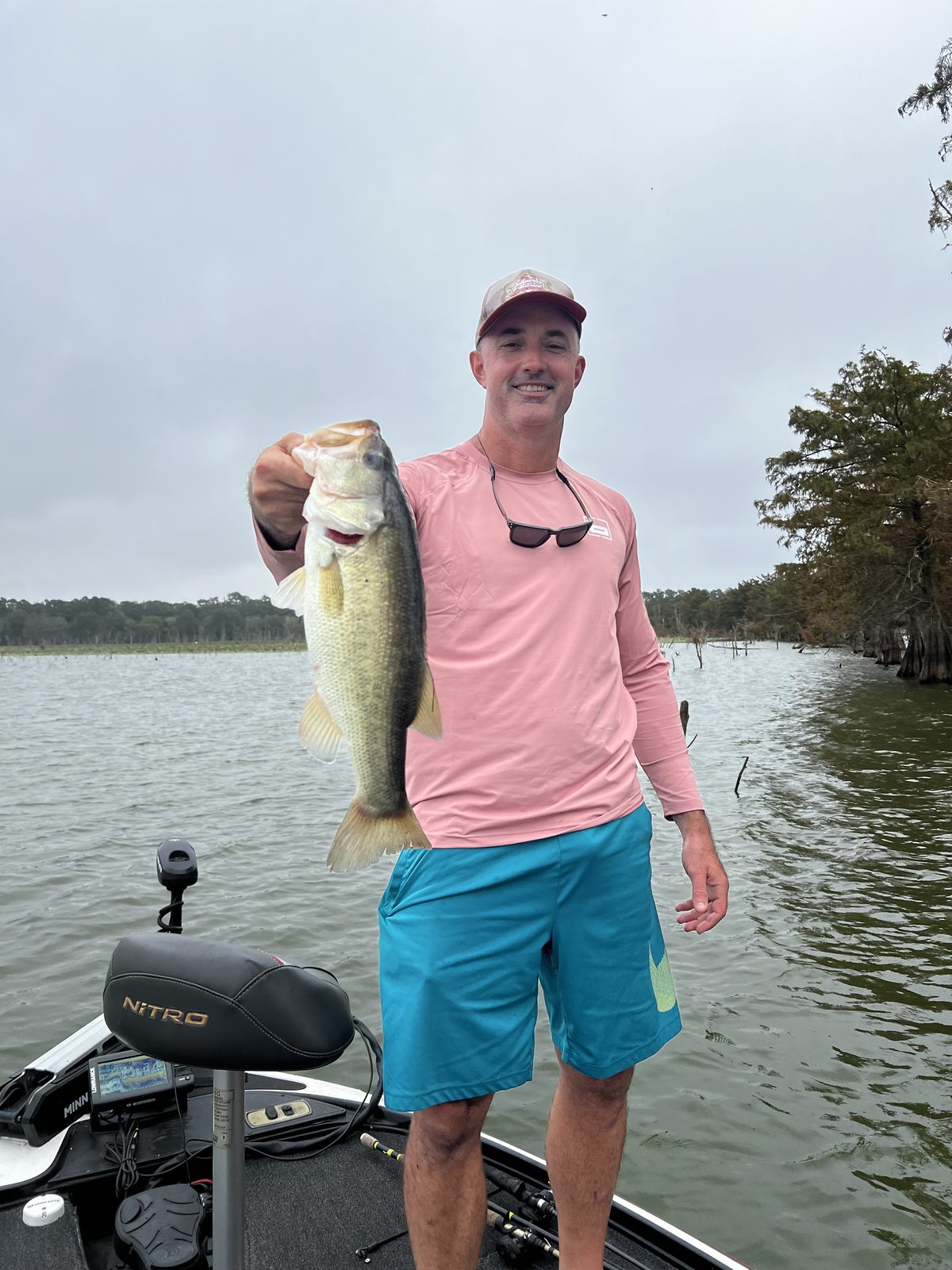 Jeremiah King on the front deck of his bass boat holding up a largemouth bass, with cypress trees and the lake behind him